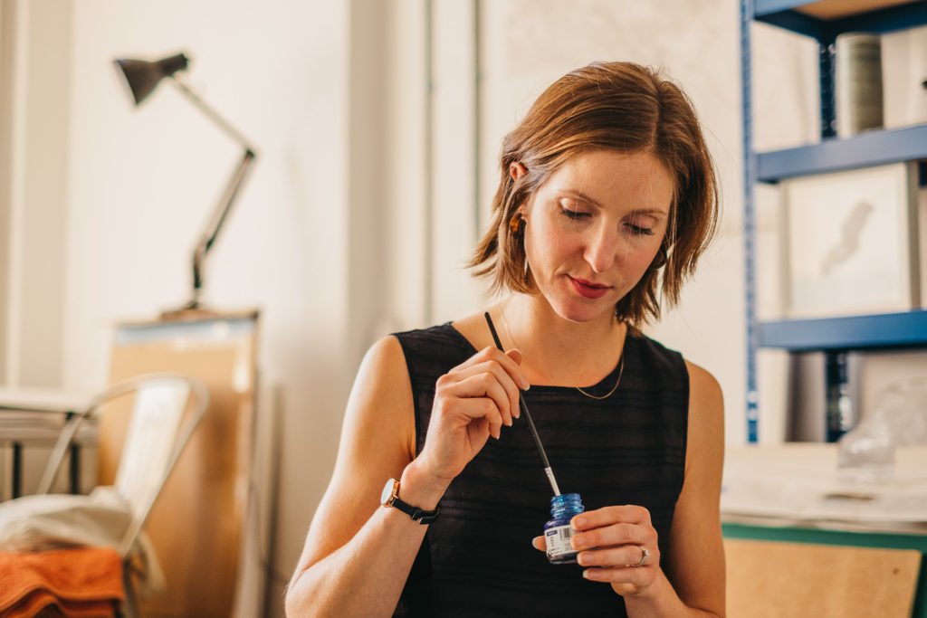 Photograph of artist Abi Spendlove dipping a brush in a pot of ink on her studio floor